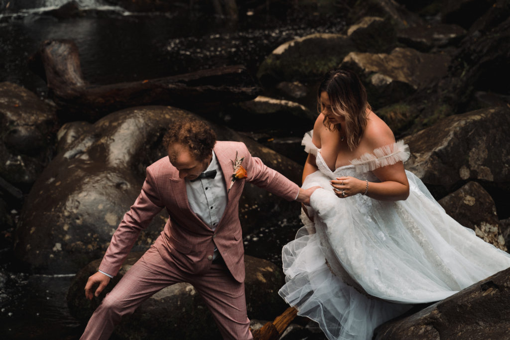 groom leads his bride over rocks at a waterfall on their wilderness elopement