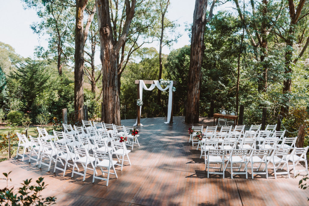 outdoor wedding ceremony location setup with a wooden arbor and white chars with green and red flowers